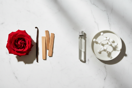 Rose bloom, vanilla pod and sandalwood beside clear vial and crystalline molecule on white marble in scientific natural light
